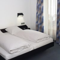 Ein schlichtes Hotelzimmer mit Doppelbett, weißen Bettdecken und blau-braunen Vorhängen. An der Wand sind zwei Leselampen angebracht., © TOMAS