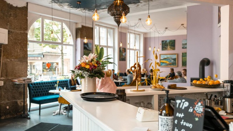 Gemütliches Café mit Blumen und Brezeln auf der Theke, blauen Sofas und Kunst an den Wänden. Große Fenster lassen viel Licht herein., © SMG, Sarah Schmid Gemütliches Café mit Blumen und Brezeln auf der Theke, blauen Sofas und Kunst an den Wänden. Große Fenster lassen viel Licht herein., © SMG, Sarah Schmid