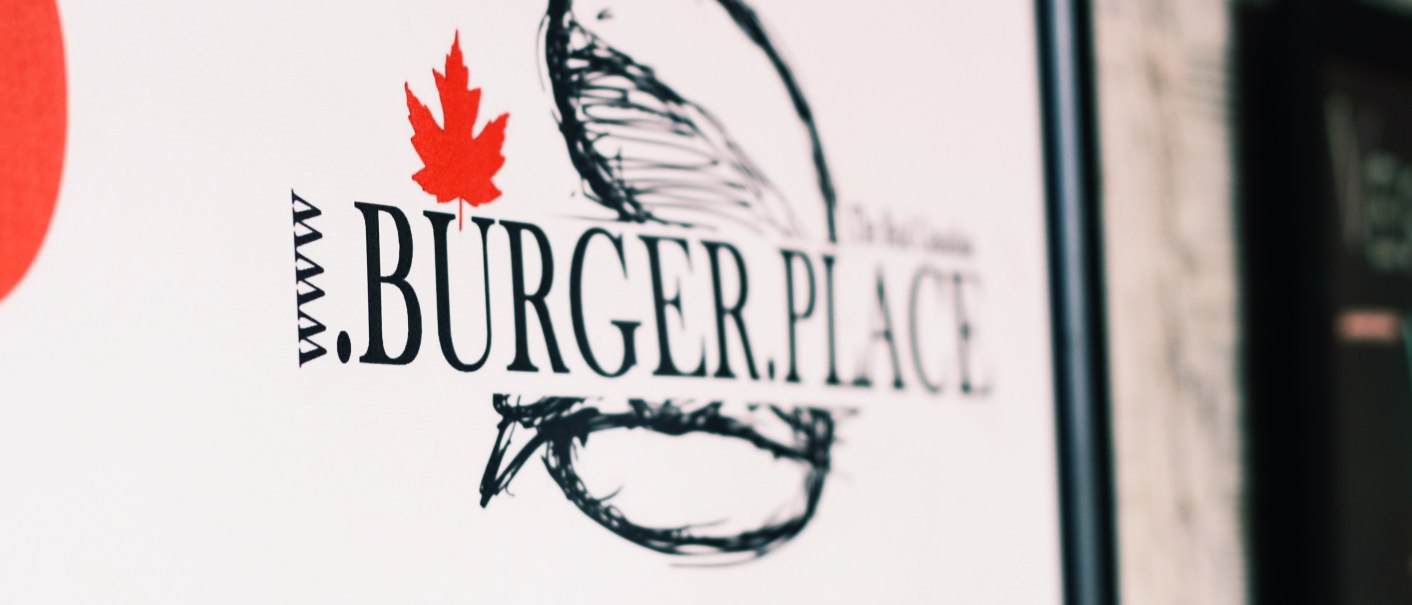 Nahaufnahme eines Schildes mit der Aufschrift "BURGER.PLACE" und einem roten Ahornblatt. Der Hintergrund ist unscharf., © SMG