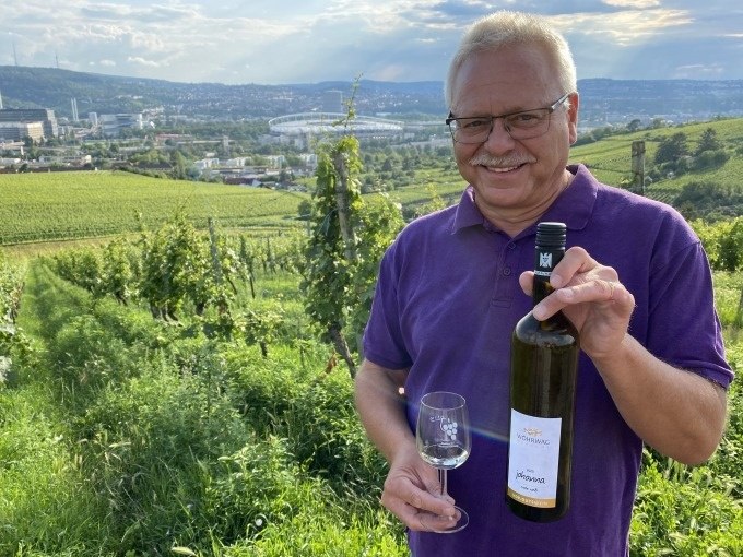 Ein Mann in einem lila Hemd hält eine Weinflasche und ein Glas in einem Weinberg. Im Hintergrund ist Stuttgart mit einem Stadion zu sehen., © Weingenuss Stuttgart