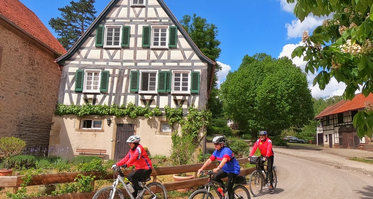 Drei Radfahrer in bunter Kleidung fahren an einem malerischen Fachwerkhaus vorbei, umgeben von grünen Bäumen und blauem Himmel., © Land der 1000 Hügel - Kraichgau-Stromberg Drei Radfahrer in bunter Kleidung fahren an einem malerischen Fachwerkhaus vorbei, umgeben von grünen Bäumen und blauem Himmel., © Land der 1000 Hügel - Kraichgau-Stromberg