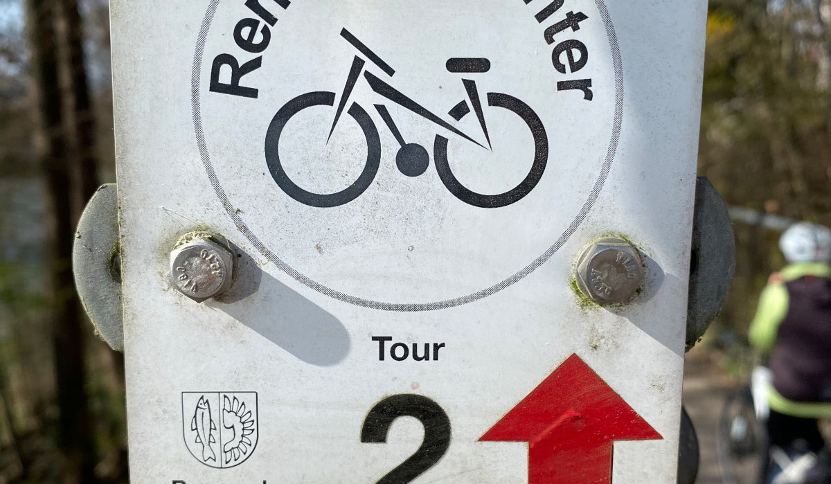 Schild mit 'Remseck-Achter Tour 2' und Fahrradsymbol an einem Pfosten, im Hintergrund ein Radfahrer auf einem Weg. Schild mit 'Remseck-Achter Tour 2' und Fahrradsymbol an einem Pfosten, im Hintergrund ein Radfahrer auf einem Weg.
