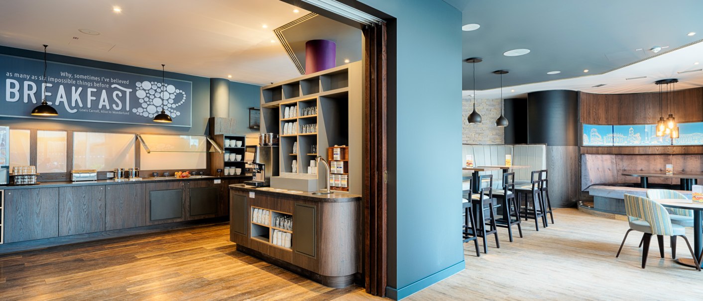 Moderner Frühstücksbereich mit Selbstbedienungstheke, Regalen und Sitzgelegenheiten. Helle Holzböden und stilvolle Beleuchtung schaffen eine einladende Atmosphäre., © Premier Inn Germany