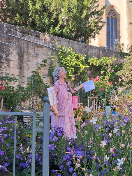 Eine als Putzfee „Frau Kunzle“ verkleidete Stadtführerin steht mit Besen, Kehrblech und pinkem Eimer in einem blühenden Garten vor einer historischen Steinmauer und einer Kirche. Sie hält ein Blatt in der Hand und erzählt während einer Stadtführung., © Stadt Leinfelden-Echterdingen | Yvonne Emhardt Eine als Putzfee „Frau Kunzle“ verkleidete Stadtführerin steht mit Besen, Kehrblech und pinkem Eimer in einem blühenden Garten vor einer historischen Steinmauer und einer Kirche. Sie hält ein Blatt in der Hand und erzählt während einer Stadtführung., © Stadt Leinfelden-Echterdingen | Yvonne Emhardt