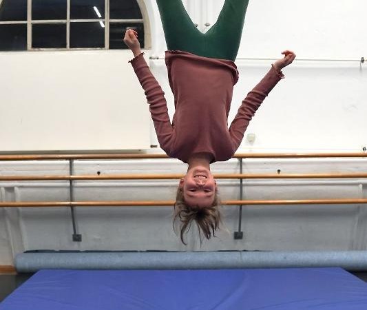 Ein Kind hängt kopfüber an einem Trapez in einem Turnraum mit blauer Matte und Fenster im Hintergrund., © Kunstschule Labyrinth Ein Kind hängt kopfüber an einem Trapez in einem Turnraum mit blauer Matte und Fenster im Hintergrund., © Kunstschule Labyrinth
