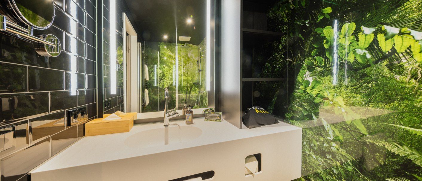 Modernes Badezimmer mit weißem Waschbecken, Spiegel und Dschungelmotiv an der Wand. Schwarze Fliesen und helle Beleuchtung ergänzen das Design., © Rioca Stuttgart Posto 4 GmbH