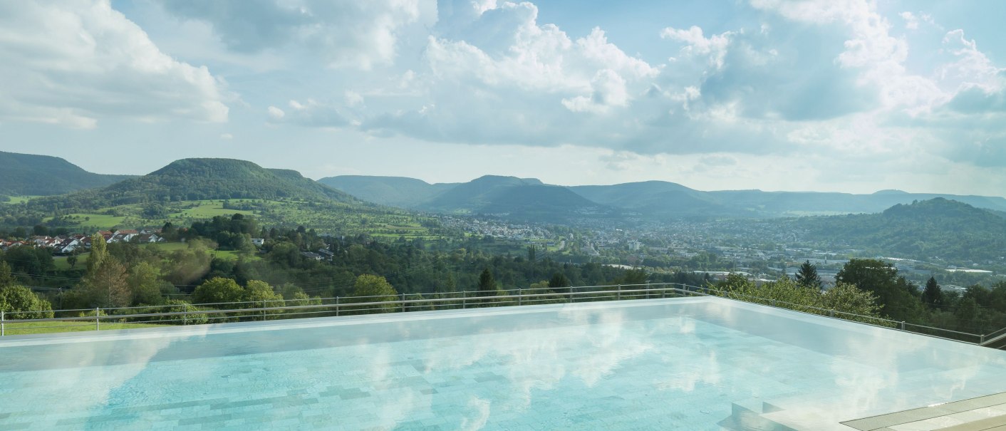 Infinity-Pool mit Blick auf grüne Hügel und eine Stadt im Tal. Der Himmel ist bewölkt, die Landschaft wirkt friedlich und einladend., © Achalm. Hotel GmbH & Co. KG Infinity-Pool mit Blick auf grüne Hügel und eine Stadt im Tal. Der Himmel ist bewölkt, die Landschaft wirkt friedlich und einladend., © Achalm. Hotel GmbH & Co. KG