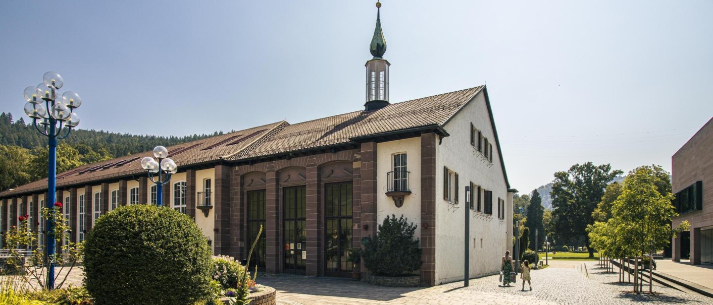 Ein historisches Geb&auml;ude in Bad Liebenzell mit einem markanten Turm und umgeben von gr&uuml;ner Landschaft. Menschen spazieren auf dem gepflasterten Weg., &copy; SMG, Sarah Schmid