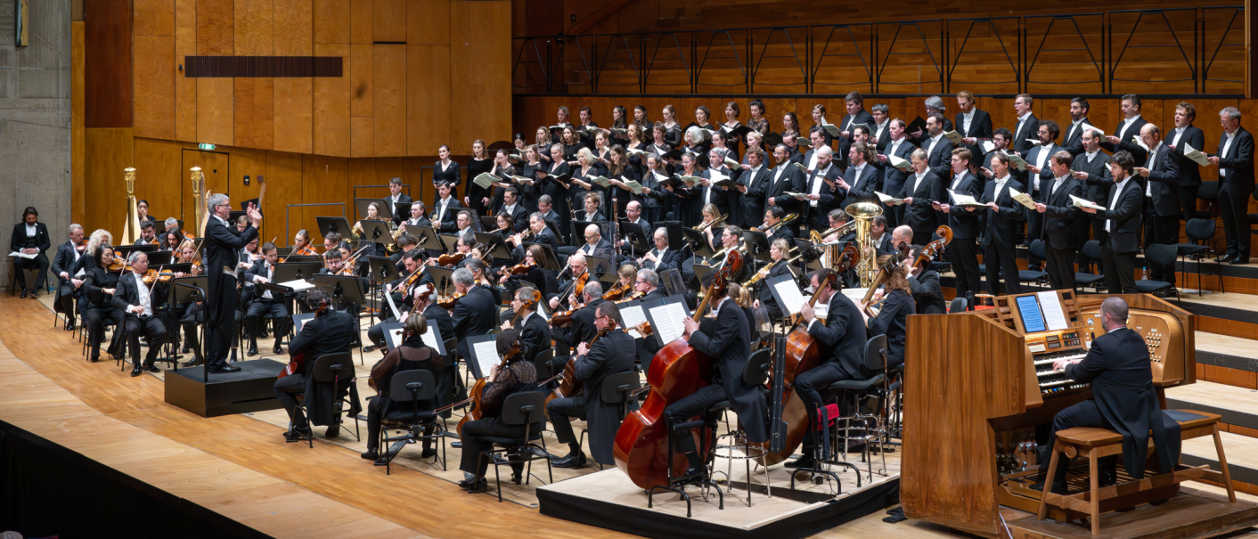 Ein gro&szlig;es Orchester und Chor f&uuml;hren in einem Konzertsaal auf. Der Dirigent steht vor den Musikern, w&auml;hrend ein Organist an der Orgel spielt., &copy; Holger Schneider