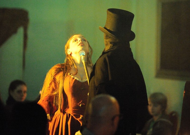 Eine Frau in einem roten Kleid steht vor einem Mann in schwarzem Mantel und Zylinder. Die Szene wirkt dramatisch und ist in warmem Licht gehalten., © Gruseldinner GmbH & Co. KG Eine Frau in einem roten Kleid steht vor einem Mann in schwarzem Mantel und Zylinder. Die Szene wirkt dramatisch und ist in warmem Licht gehalten., © Gruseldinner GmbH & Co. KG