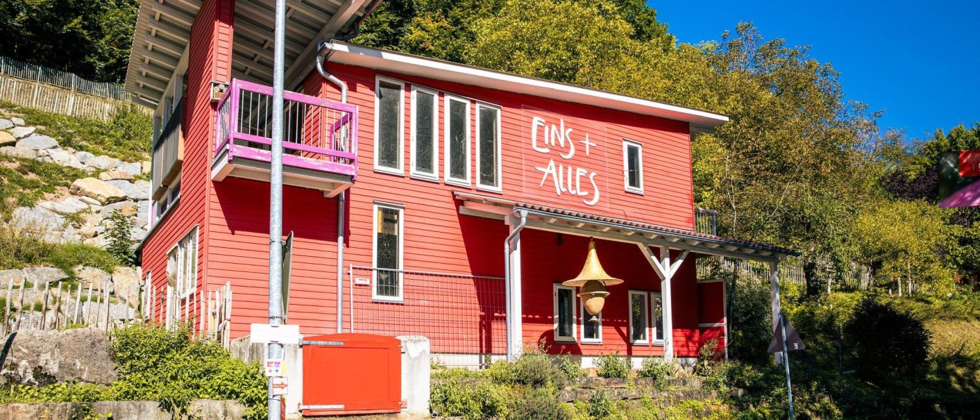 Ein rotes Geb&auml;ude mit der Aufschrift 'Eins + Alles' steht in einer gr&uuml;nen, bewaldeten Umgebung. Es hat gro&szlig;e Fenster und einen kleinen Balkon., &copy; Stuttgart-Marketing GmbH, Sarah Schmid