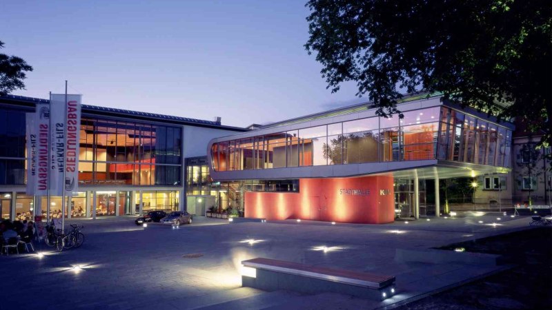 Die Stadthalle K3N in N&uuml;rtingen bei D&auml;mmerung, modern beleuchtet mit Glasfassade und rotem Akzent. Fahrr&auml;der und Autos sind im Vordergrund sichtbar., &copy; Peschke