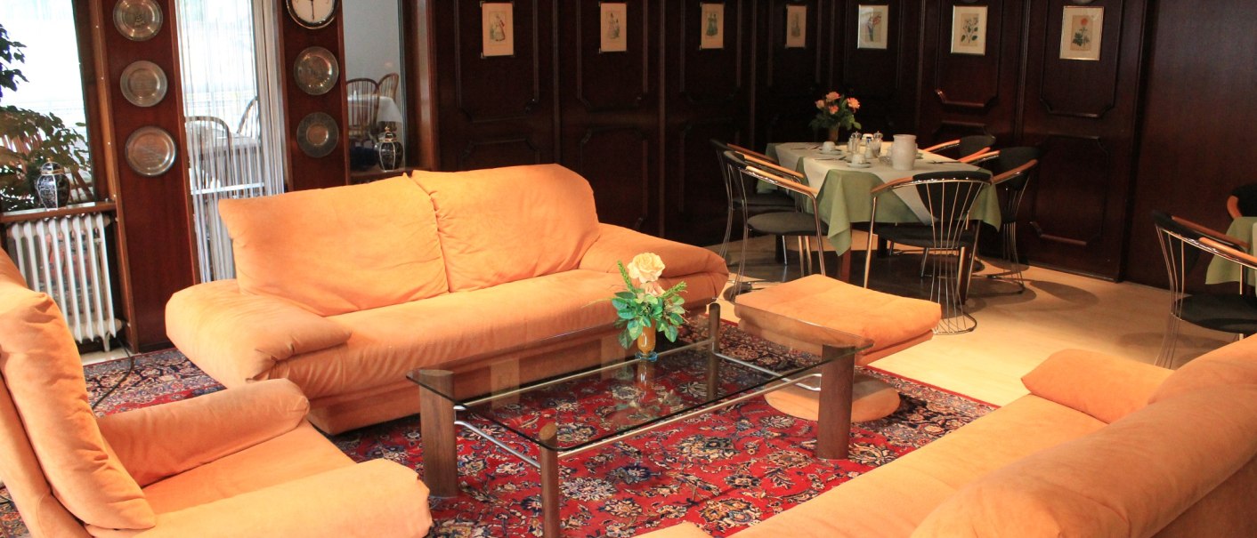 Gem&uuml;tlicher Loungebereich im Hotel mit orangen Sofas, Glastisch und rotem Teppich. Im Hintergrund sind gedeckte Tische und Holzw&auml;nde mit Dekoration., &copy; Hotel Spahr