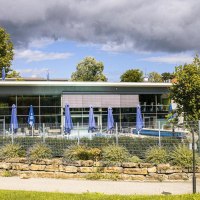 Au&szlig;enansicht der Mineraltherme Bad Boll, umgeben von B&auml;umen. Blaue Sonnenschirme stehen auf der Terrasse, ein Zaun umgibt das Gel&auml;nde., &copy; Stuttgart-Marketing GmbH, Sarah Schmid
