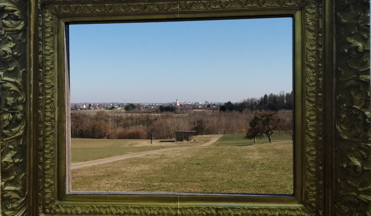 Ein goldener Rahmen umrahmt eine Landschaft mit Feldern, einem Weg und einem Dorf im Hintergrund unter klarem Himmel., © Foto: Cornelia Steinbach Ein goldener Rahmen umrahmt eine Landschaft mit Feldern, einem Weg und einem Dorf im Hintergrund unter klarem Himmel., © Foto: Cornelia Steinbach