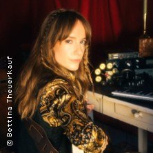 Frau mit langen Haaren und gemustertem Oberteil sitzt an einem Tisch mit Musikequipment vor rotem Vorhang., © links im Bild