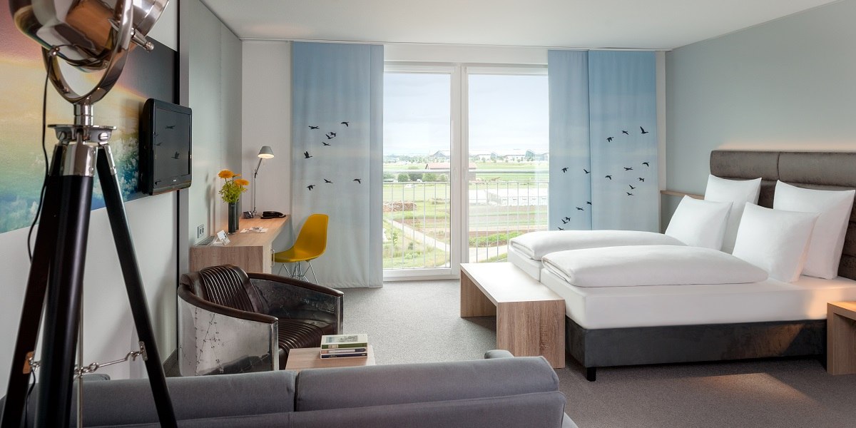 Modernes Hotelzimmer mit großem Bett, Schreibtisch, Sessel und Balkon mit Aussicht auf Landschaft. Dekor mit blauen Vorhängen und Vogelmotiven., © Essential by Dorint
