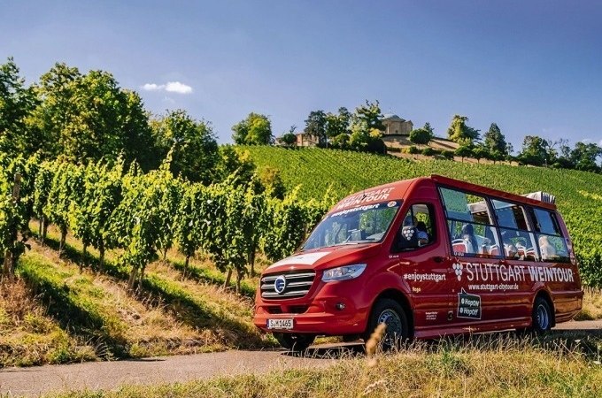 Ein roter Tourbus mit der Aufschrift 'Stuttgart Weintour' fährt durch grüne Weinberge unter blauem Himmel., © SMG, Thomas Niedermüller