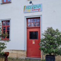 Der Eingangsbereich der Abessina Café Lounge mit roter Tür und Fenstern, flankiert von zwei Pflanzen in Kübeln., © cAbessina Der Eingangsbereich der Abessina Café Lounge mit roter Tür und Fenstern, flankiert von zwei Pflanzen in Kübeln., © cAbessina