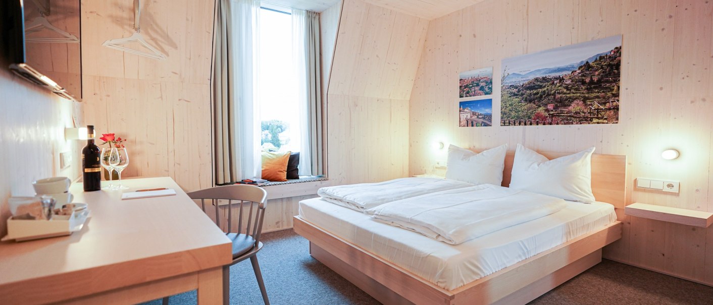 Modernes Hotelzimmer mit Holzwänden, Doppelbett, Schreibtisch und Fensterbank. An der Wand hängen Bilder von Landschaften., © Hotel Bergamo