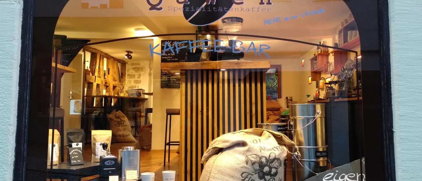 Schaufenster der Kaffeerösterei Qaweh mit Kaffeesäcken, Produkten und einer Kaffeemaschine. Schriftzug 'Kaffeebar' und 'Spezialitätenkaffee' sichtbar., © Matthias Gohl Schaufenster der Kaffeerösterei Qaweh mit Kaffeesäcken, Produkten und einer Kaffeemaschine. Schriftzug 'Kaffeebar' und 'Spezialitätenkaffee' sichtbar., © Matthias Gohl