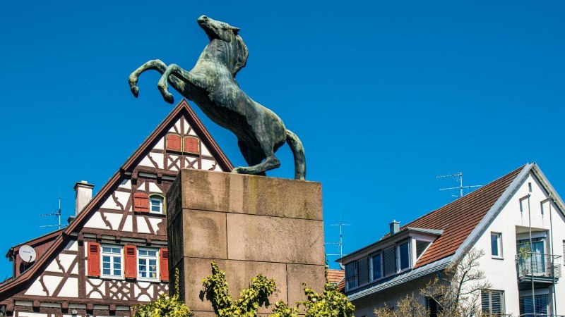 Eine Pferdestatue auf einem Sockel vor traditionellen Fachwerkhäusern in Kirchheim unter Teck, unter klarem, blauem Himmel., © Stuttgart-Marketing GmbH, Sarah Schmid
