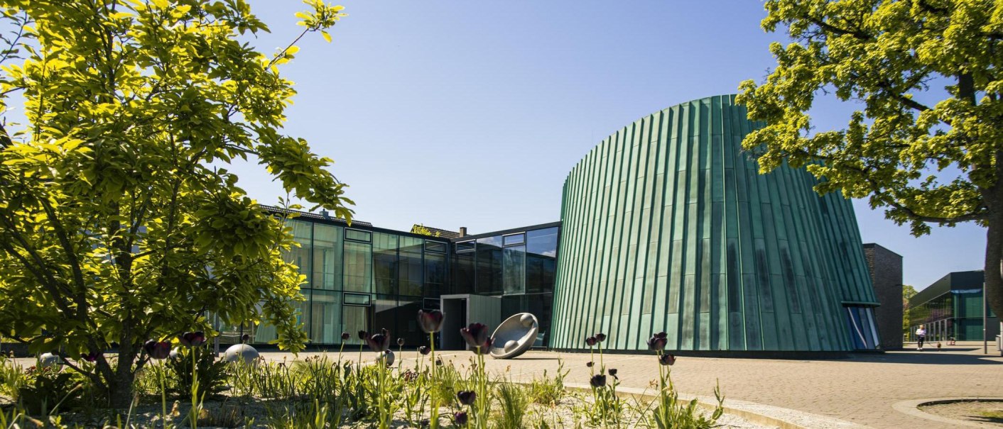 Die Musikschule Fellbach zeigt moderne Architektur mit einem grünen, konischen Gebäude und Glasfassaden, umgeben von Bäumen und Blumen., © SMG, Sarah Schmid Die Musikschule Fellbach zeigt moderne Architektur mit einem grünen, konischen Gebäude und Glasfassaden, umgeben von Bäumen und Blumen., © SMG, Sarah Schmid