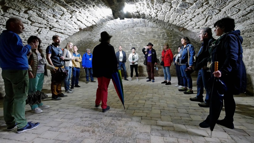 Menschen stehen in einem Gewölbekeller aus Stein, hören einer Person mit Hut zu. Der Raum ist mit Kopfsteinpflaster ausgelegt., © Clemens Scholl