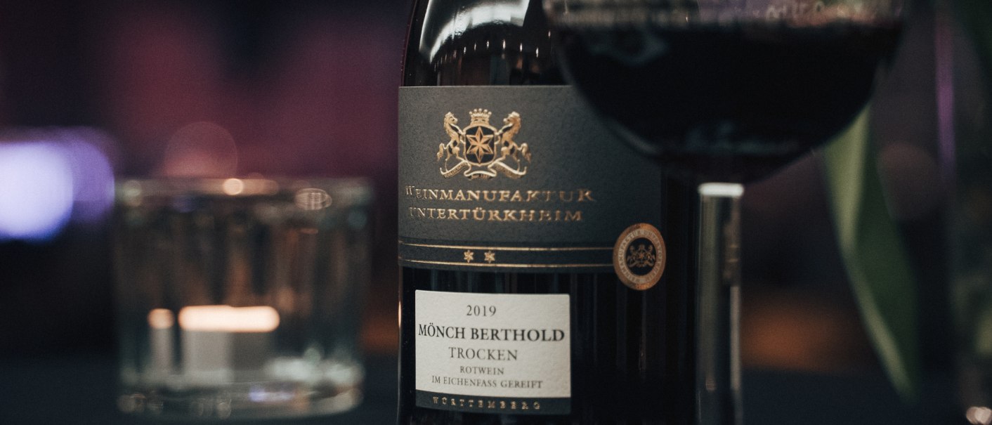 Nahaufnahme einer Weinflasche "Mönch Berthold" von 2019, trocken, mit einem Glas Rotwein daneben. Elegantes Etikett mit goldener Schrift., © Weinmanufaktur Untertürkheim