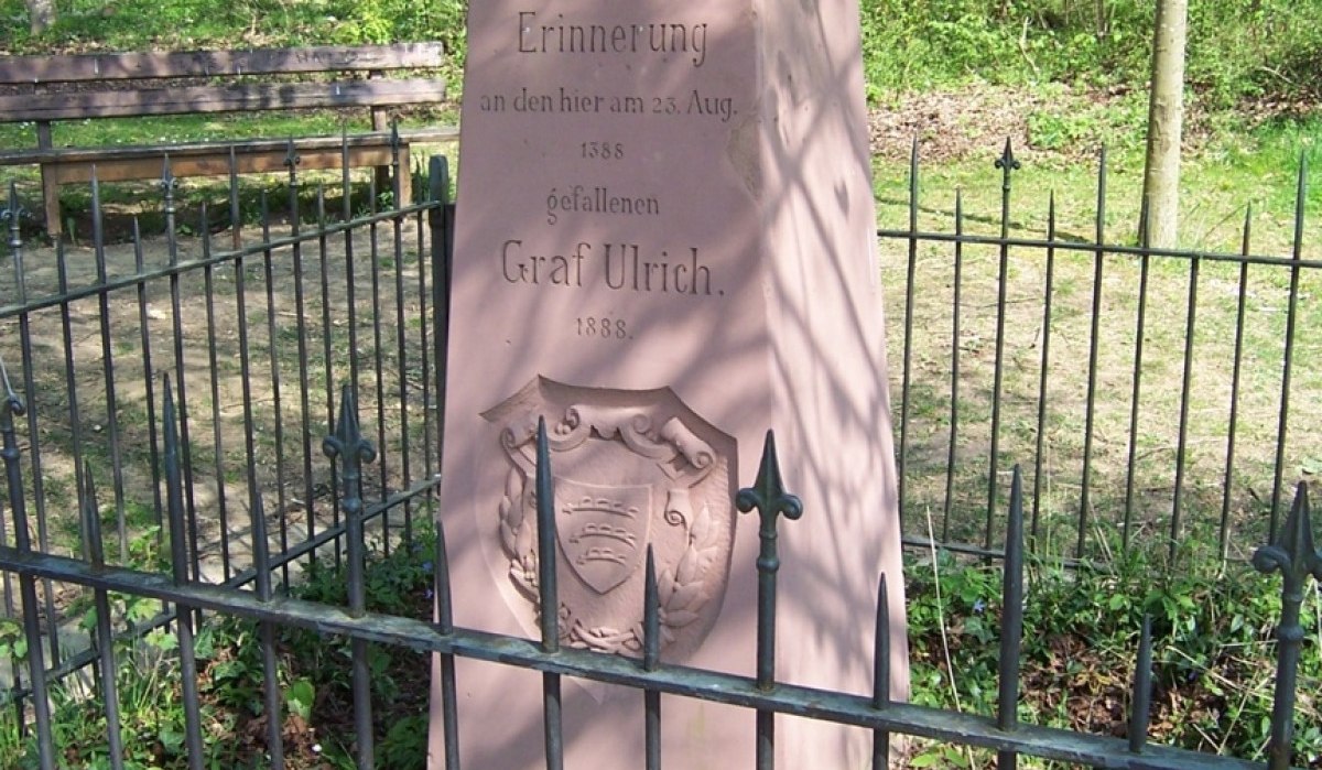 Ein Gedenkstein für Graf Ulrich, umgeben von einem Zaun, steht in einem Wald. Der Stein erinnert an den Tod von Graf Ulrich im Jahr 1388., © Natur.Nah. Schönbuch & Heckengäu Ein Gedenkstein für Graf Ulrich, umgeben von einem Zaun, steht in einem Wald. Der Stein erinnert an den Tod von Graf Ulrich im Jahr 1388., © Natur.Nah. Schönbuch & Heckengäu
