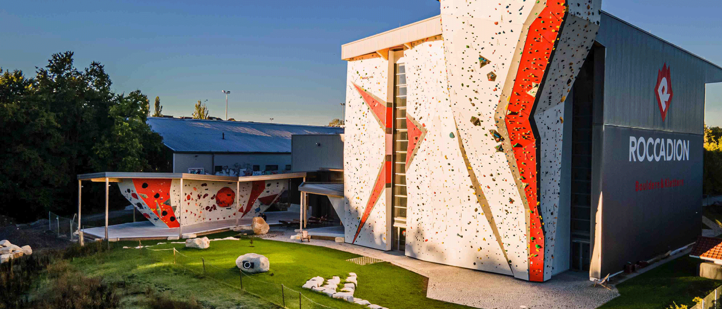 Kletterhalle mit hohen Kletterwänden und Boulderbereich im Freien, umgeben von Bäumen und Wiese, bei Sonnenuntergang., © Roccadion, Böblingen Kletterhalle mit hohen Kletterwänden und Boulderbereich im Freien, umgeben von Bäumen und Wiese, bei Sonnenuntergang., © Roccadion, Böblingen