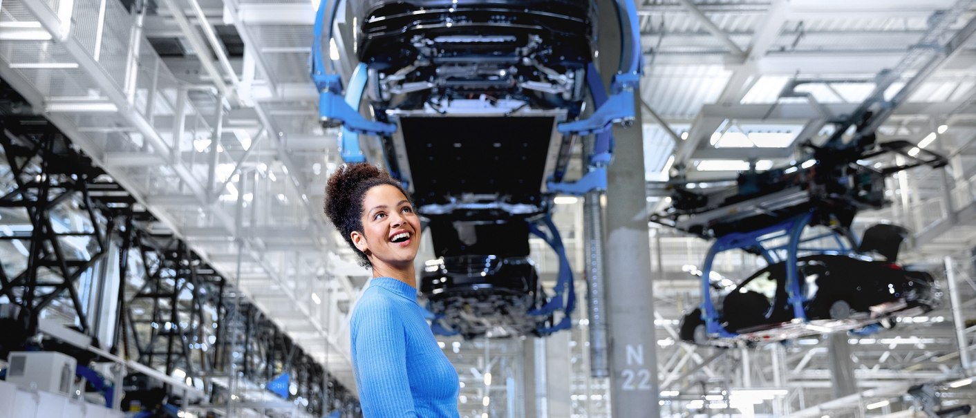 Frau in blauen Pullover in moderner Autofabrik mit hängenden Autokarosserien im Hintergrund., © Mercedes-Benz AG Frau in blauen Pullover in moderner Autofabrik mit hängenden Autokarosserien im Hintergrund., © Mercedes-Benz AG