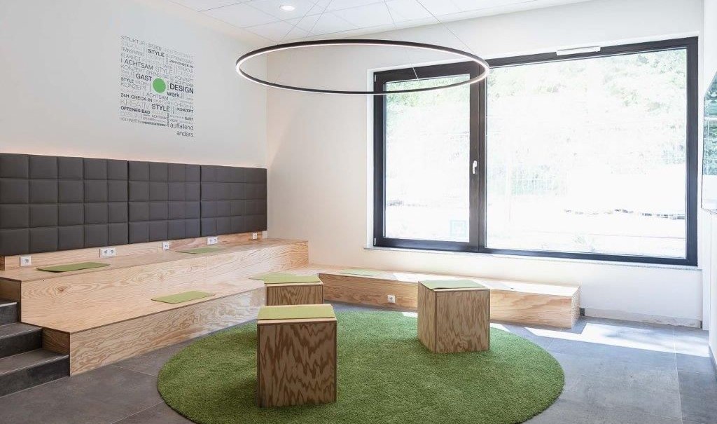 Moderne Lobby mit Holzbänken, grünen Sitzkissen und einem großen Fenster. Ein runder Teppich und eine kreisförmige Lampe ergänzen das Design., © a2 HOTELS am Quadrium