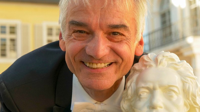 Ein lächelnder Mann in einem Anzug hält eine Beethoven-Büste vor einem historischen Gebäude mit gelber Fassade., © Stadtverwaltung Schorndorf