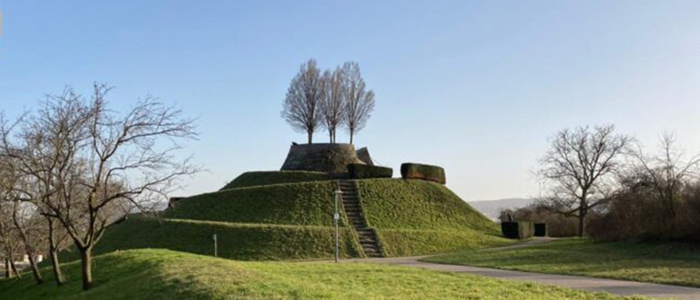 Eine grasbewachsene Erhebung mit einer Treppe und Bäumen auf der Spitze, umgeben von kahlen Bäumen, unter einem klaren blauen Himmel., © SMG Eine grasbewachsene Erhebung mit einer Treppe und Bäumen auf der Spitze, umgeben von kahlen Bäumen, unter einem klaren blauen Himmel., © SMG