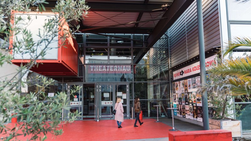Eingang des Theaterhauses Stuttgart mit rotem Boden, Glasfassade und Plakaten. Zwei Personen betreten das Geb&auml;ude. Pflanzen und moderne Architektur sind sichtbar., &copy; Stuttgart-Marketing GmbH, wpsteinheisser