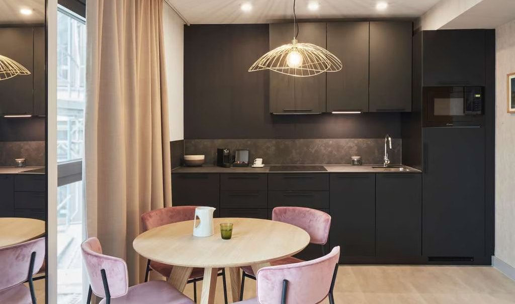 Moderne Küche mit schwarzer Einbauküche, rundem Holztisch und rosa Stühlen. Große Fenster und stilvolle Beleuchtung., © Adagio Moderne Küche mit schwarzer Einbauküche, rundem Holztisch und rosa Stühlen. Große Fenster und stilvolle Beleuchtung., © Adagio