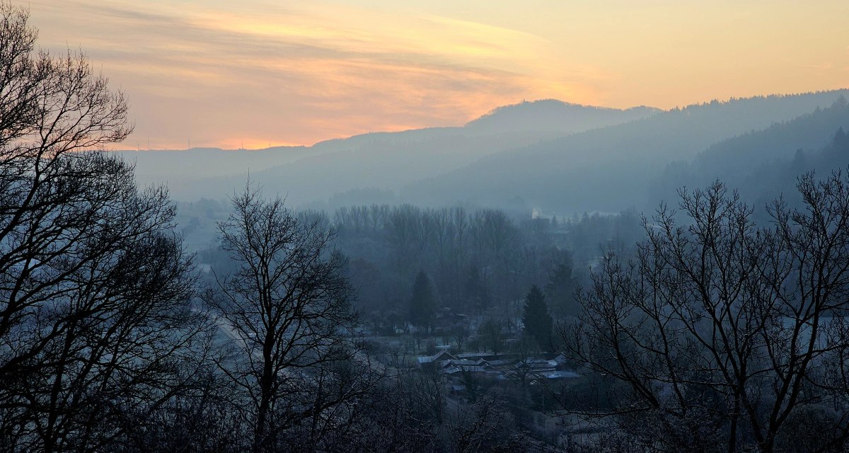 Sonnenuntergang über nebliger Landschaft, Bäume im Vordergrund, Hügel im Hintergrund. Der Himmel ist in warmen Farben gefärbt. Sonnenuntergang über nebliger Landschaft, Bäume im Vordergrund, Hügel im Hintergrund. Der Himmel ist in warmen Farben gefärbt.
