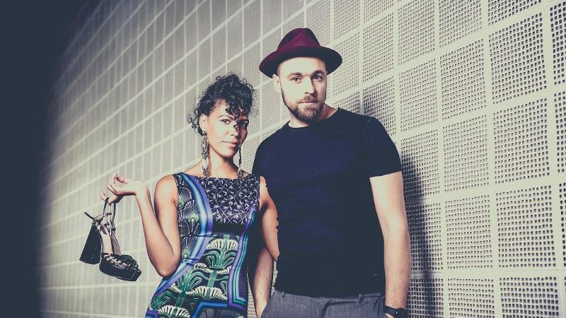 Eine Frau in einem bunten Kleid hält Schuhe, neben ihr steht ein Mann mit Hut vor einer strukturierten Wand., © Backnang Eine Frau in einem bunten Kleid hält Schuhe, neben ihr steht ein Mann mit Hut vor einer strukturierten Wand., © Backnang