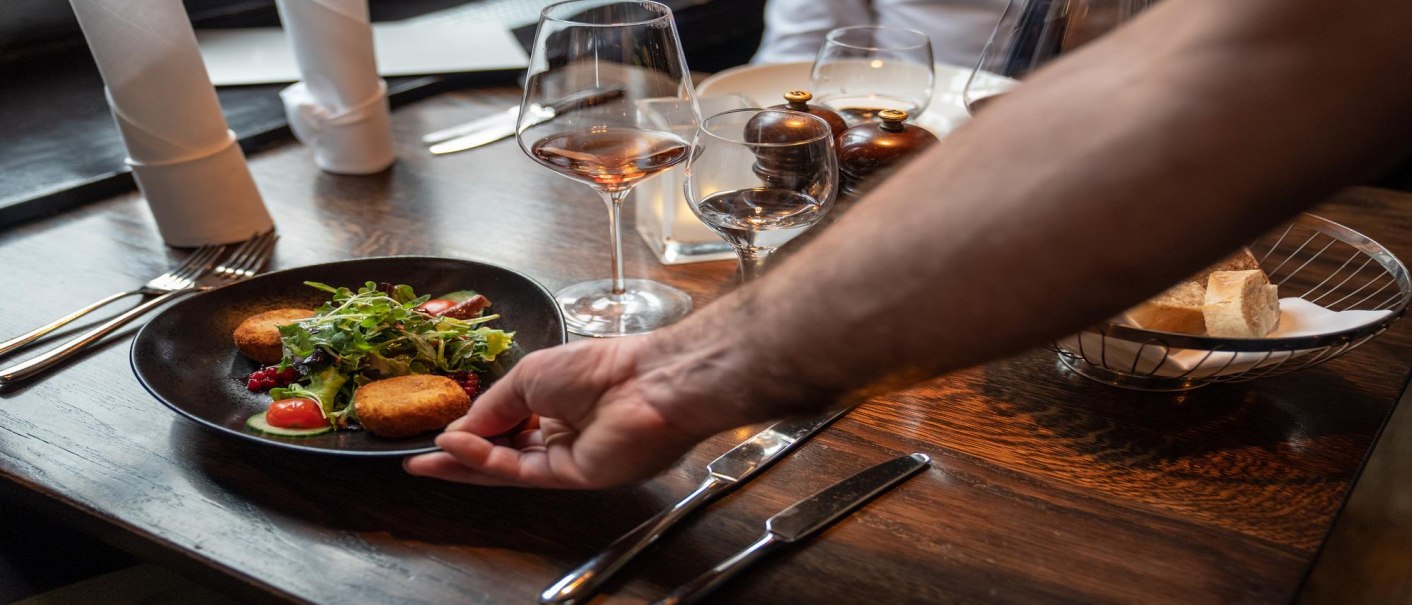 Ein Teller mit Salat und Kroketten wird auf einem gedeckten Tisch in einem Restaurant serviert. Im Hintergrund stehen Weingläser und ein Brotkorb., © Stuttgart-Marketing GmbH, Martina Denker