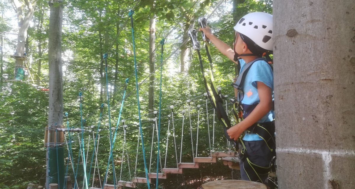 Ein Kind mit Helm und Kletterausrüstung steht in einem Kletterwald und greift nach einem Seil. Im Hintergrund sind Bäume und ein Kletterparcours zu sehen., © Kulturamt Plochingen Ein Kind mit Helm und Kletterausrüstung steht in einem Kletterwald und greift nach einem Seil. Im Hintergrund sind Bäume und ein Kletterparcours zu sehen., © Kulturamt Plochingen