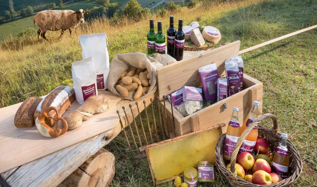 Regionale Produkte wie Brot, Wein, Äpfel und Honig auf einem Tisch im Freien. Ein Schaf steht im Hintergrund auf einer Wiese.