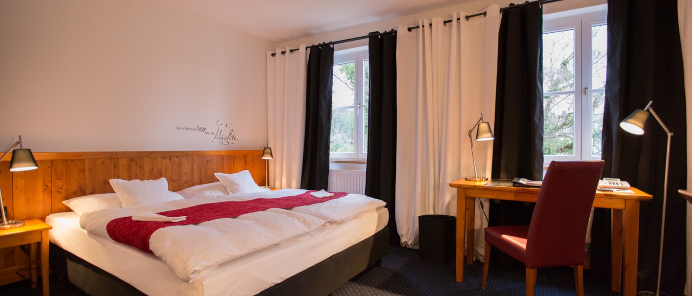 Ein gem&uuml;tliches Hotelzimmer mit Doppelbett, rotem &Uuml;berwurf, Schreibtisch und Stuhl. Gro&szlig;e Fenster mit schwarzen und wei&szlig;en Vorh&auml;ngen., &copy; Hotel B&uuml;rkle