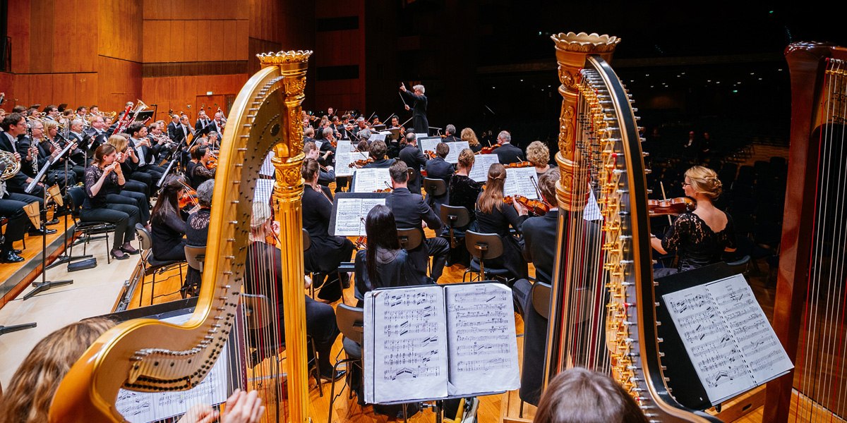 Ein Orchester spielt auf einer Bühne, mit zwei Harfen im Vordergrund. Der Dirigent steht vor den Musikern, die Notenblätter lesen., © Stuttgarter Philharmoniker
