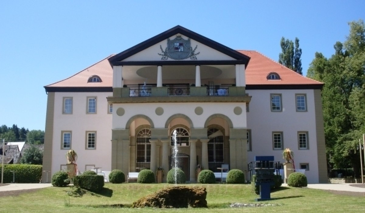 Schloss Dätzingen mit symmetrischer Fassade, rotem Dach und Säulen. Im Vordergrund ein gepflegter Garten mit Springbrunnen., © Gemeinde Grafenau
