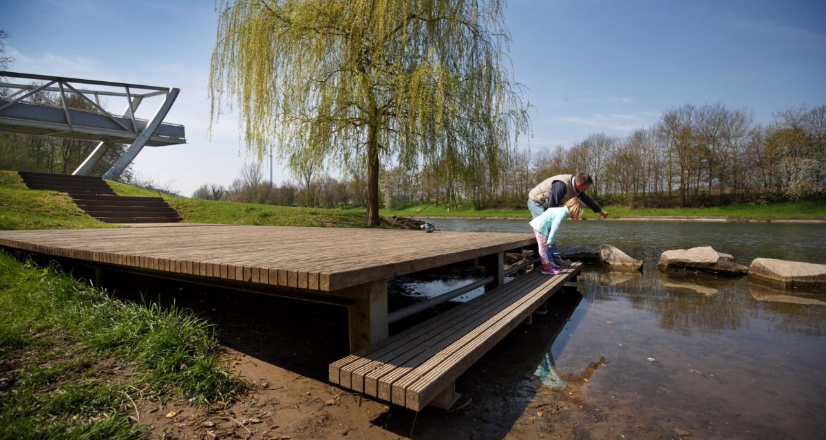 Ein Mann und ein Kind auf einem Holzsteg am Flussufer, umgeben von Natur. Eine moderne Brücke und ein Baum im Hintergrund., © Stoppel Ein Mann und ein Kind auf einem Holzsteg am Flussufer, umgeben von Natur. Eine moderne Brücke und ein Baum im Hintergrund., © Stoppel