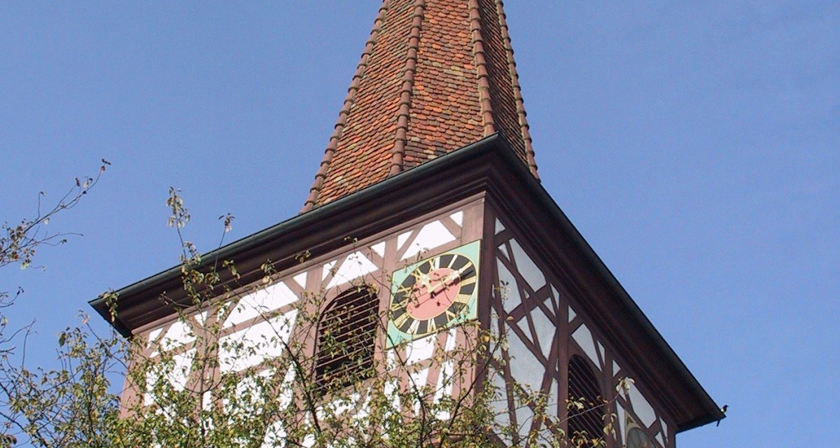 Ein Kirchturm mit Fachwerk und Uhr ragt über Bäume in den blauen Himmel. Der Turm hat ein spitzes Dach mit Wetterhahn., © Natur.Nah. Schönbuch & Heckengäu Ein Kirchturm mit Fachwerk und Uhr ragt über Bäume in den blauen Himmel. Der Turm hat ein spitzes Dach mit Wetterhahn., © Natur.Nah. Schönbuch & Heckengäu