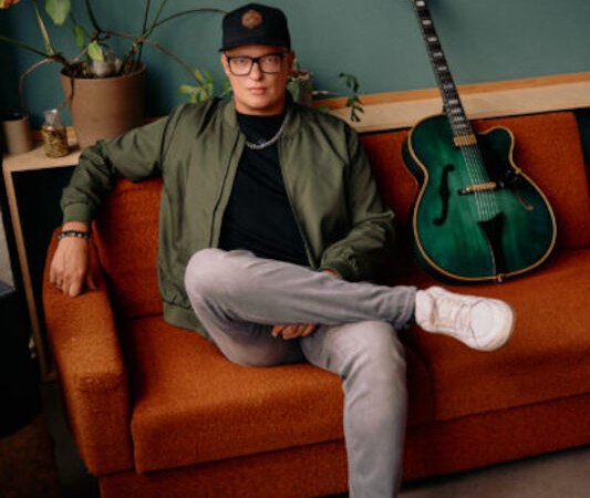 Person mit Brille und Mütze sitzt lässig auf einem orangefarbenen Sofa, eine grüne Gitarre lehnt daneben an der Wand., © Jazzclub Schwäbisch Hall e.V.