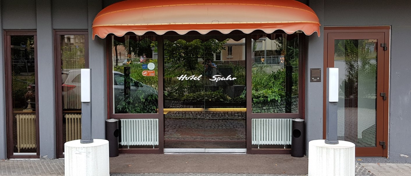 Eingang des Hotel Spahr mit Glasfront, orangefarbenem Vordach und Pflanzen im Hintergrund. Zwei wei&szlig;e S&auml;ulen stehen vor der T&uuml;r., &copy; Hotel Spahr