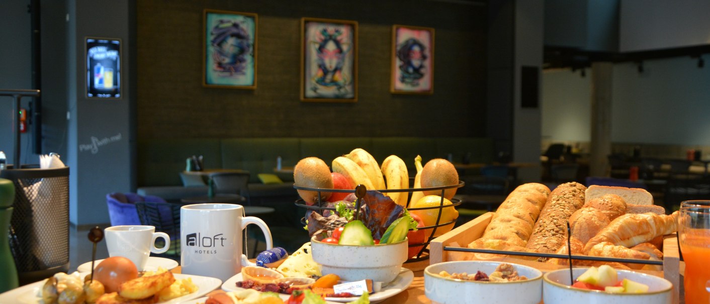 Ein &uuml;ppiges Fr&uuml;hst&uuml;cksbuffet in einem Hotelrestaurant mit Brot, Obst, M&uuml;sli und Getr&auml;nken. Im Hintergrund sind moderne Kunstwerke zu sehen., &copy; Aloft Stuttgart